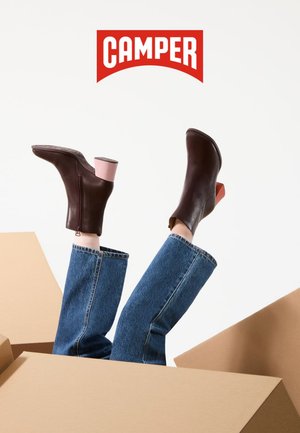 Braune Leder-Stiefeletten mit einem rosa Blockabsatz und sichtbarem Reißverschlussdetail. Kombiniert mit hellblauen Jeans mit weitem Bein, Füße ragen aus einem Karton heraus.