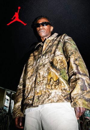 Man wearing camo jacket and sunglasses stands against dark sky with a red Air Jordan logo above left shoulder.