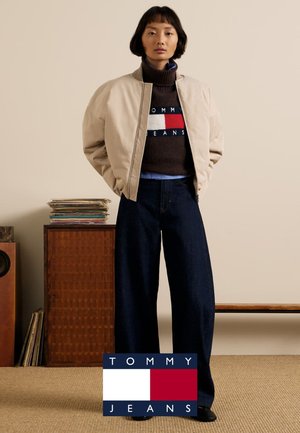 Beżowa kurtka typu bomber na brązowym swetrze z niebieskimi akcentami, w połączeniu z ciemnymi, szerokimi jeansami. Na neutralnym tle.