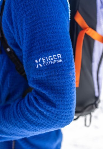 Blaues Fleecejackenärmel mit "Eiger Extreme"-Logo, getragen von einer Person, die im Freien in einer schneebedeckten Umgebung einen schwarz-orangen Rucksackgurt trägt.