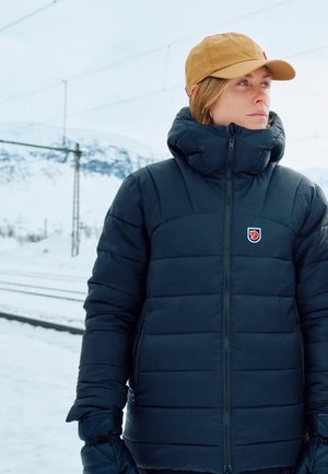Schwarze Pufferjacke mit gestepptem Muster, hohem Kragen und Reißverschluss. Mit einem Markenemblem auf der Brust. Getragen mit einer tanfarbenen Mütze und Handschuhen.