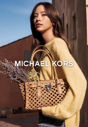 Vrouw in gele trui met gevlochten tan kleurige Michael Kors handtas met bloemen, staand buiten tegen een gebouw en een heldere blauwe lucht.