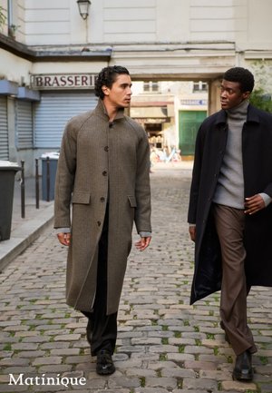 Zwei Männer gehen auf einer Kopfsteinpflasterstraße. Der eine trägt einen braunen Hahnentritt-Mantel, der andere einen schwarzen Überwurf, beide kombiniert mit dunklen Hosen und Schuhen.