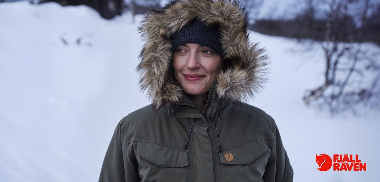 Grön parkas med pälsfodrad huva, innehållande två bröstfickor, dragkedjestängning och en logotypbadge. Snöig bakgrund förstärker den utomhusmiljön.