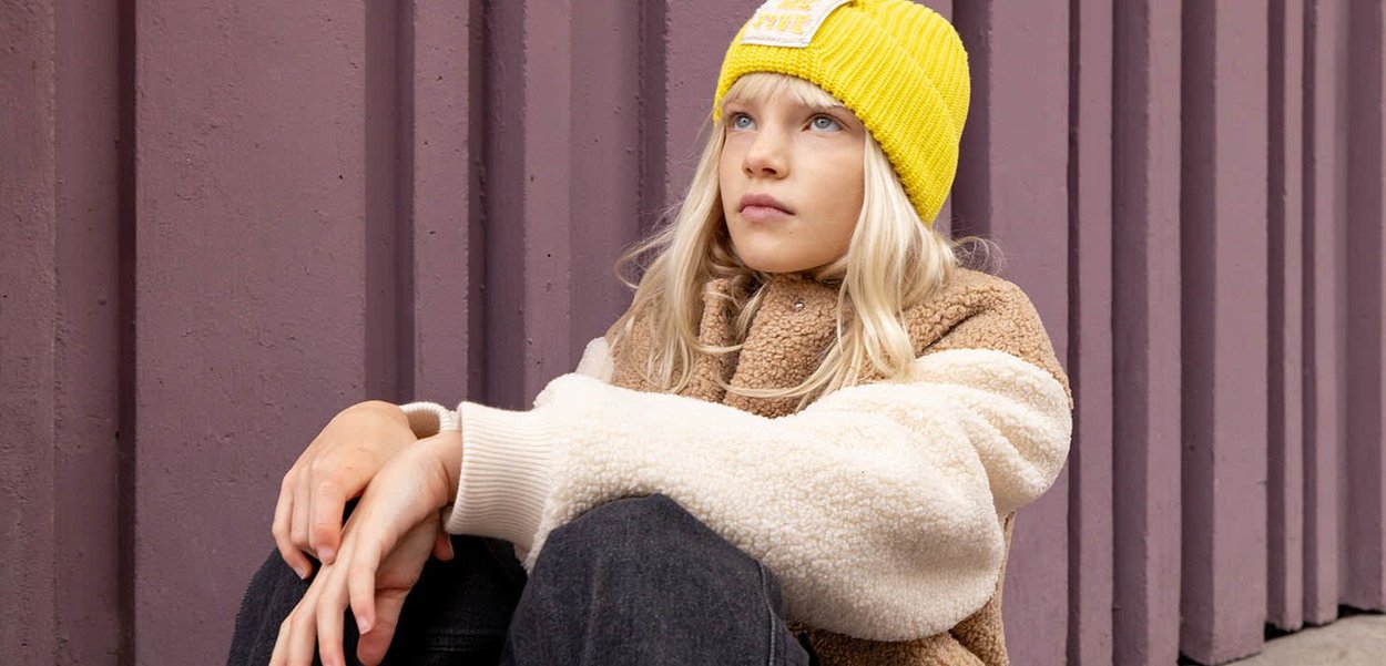 Bonnet en tricot jaune, veste en polaire beige et marron, jeans sombres. Sujet assis contre un mur texturé violet, les bras reposant sur les genoux.