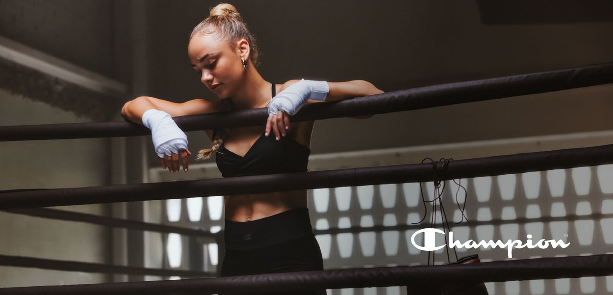 Crop top atletico nero e leggings con dettagli del logo, mani avvolte, in piedi vicino a guanti da boxe marroni in un ambiente di palestra.