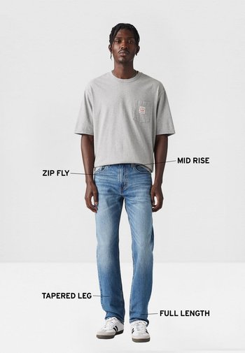 Camiseta de algodón gris con bolsillo en el pecho, vaqueros azules de tiro medio con cremallera, piernas ajustadas y diseño de largo completo, combinados con zapatillas blancas.