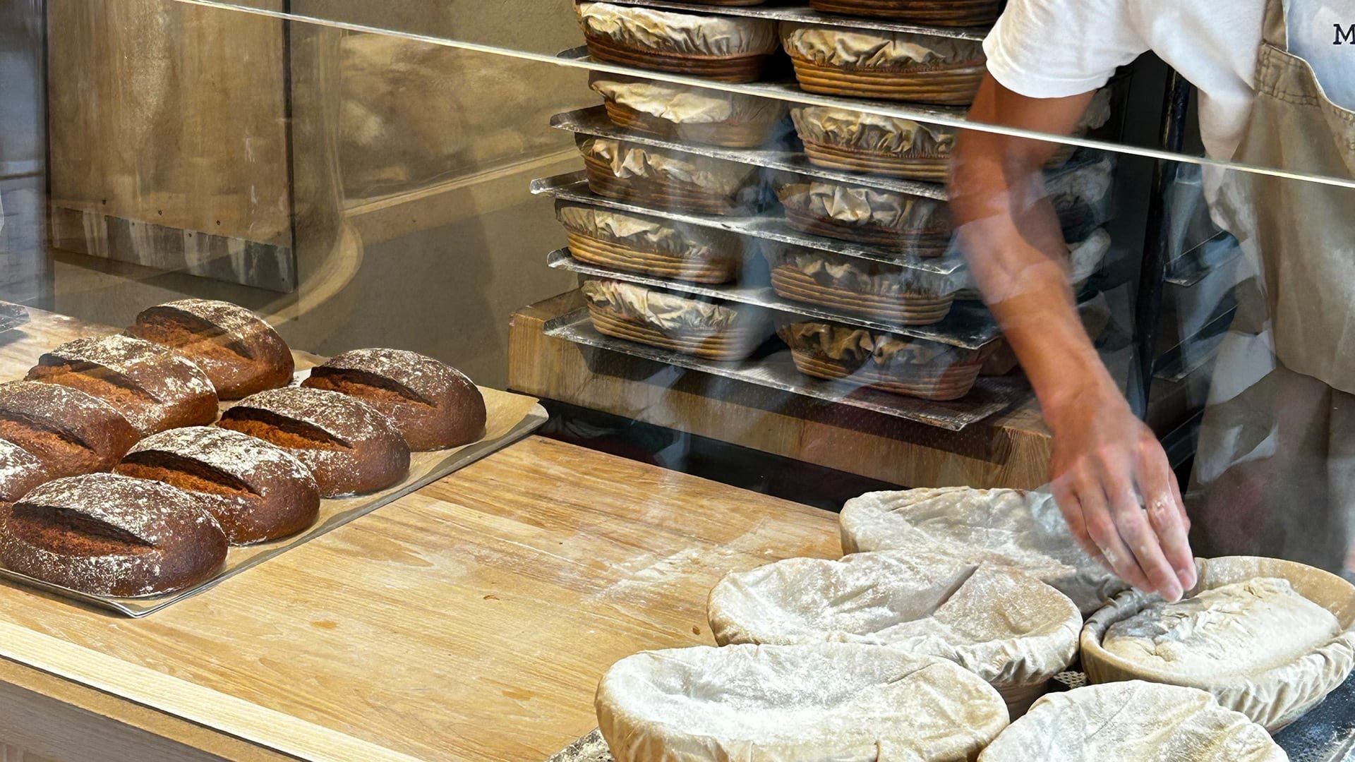 Ambachtelijke bruinbroden met een meeldusting op een houten oppervlak; ronde, rustieke vorm. Deeg in manden gestapeld aan de achterkant.