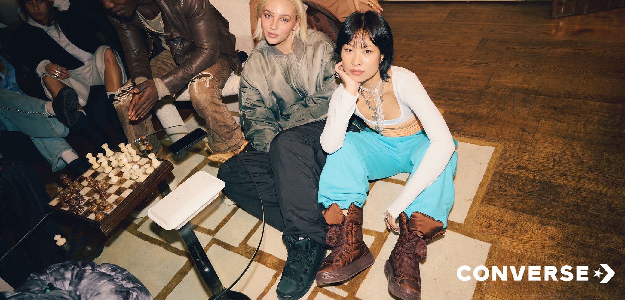 Dos personas están sentadas en un suelo de madera junto a un tablero de ajedrez. Una lleva botas marrones y pantalones azules holgados; la otra viste una chaqueta bomber verde.