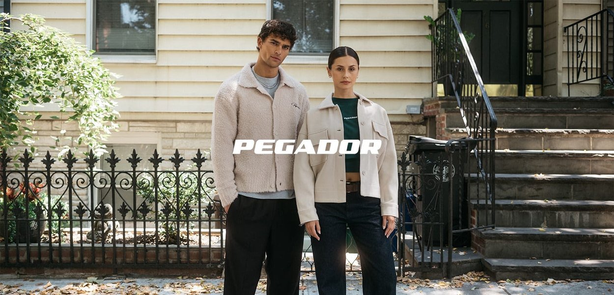Two models stand outside. The man wears a beige fleece jacket, and the woman wears a light beige overshirt. Both have dark pants.
