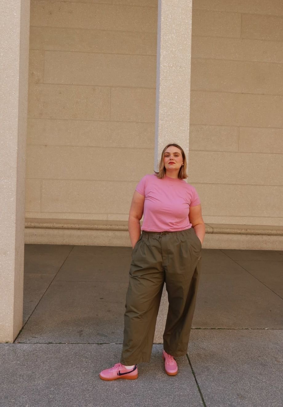 Pink short-sleeve shirt paired with loose-fitting olive green trousers and pink Nike shoes. Standing against a light-colored wall.