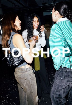 Drei Frauen lächeln und halten Getränke, vertieft in ein lebhaftes Gespräch in einer schummrig beleuchteten Gesellschaftsumgebung mit dem Text "TOPSHOP" eingeblendet.