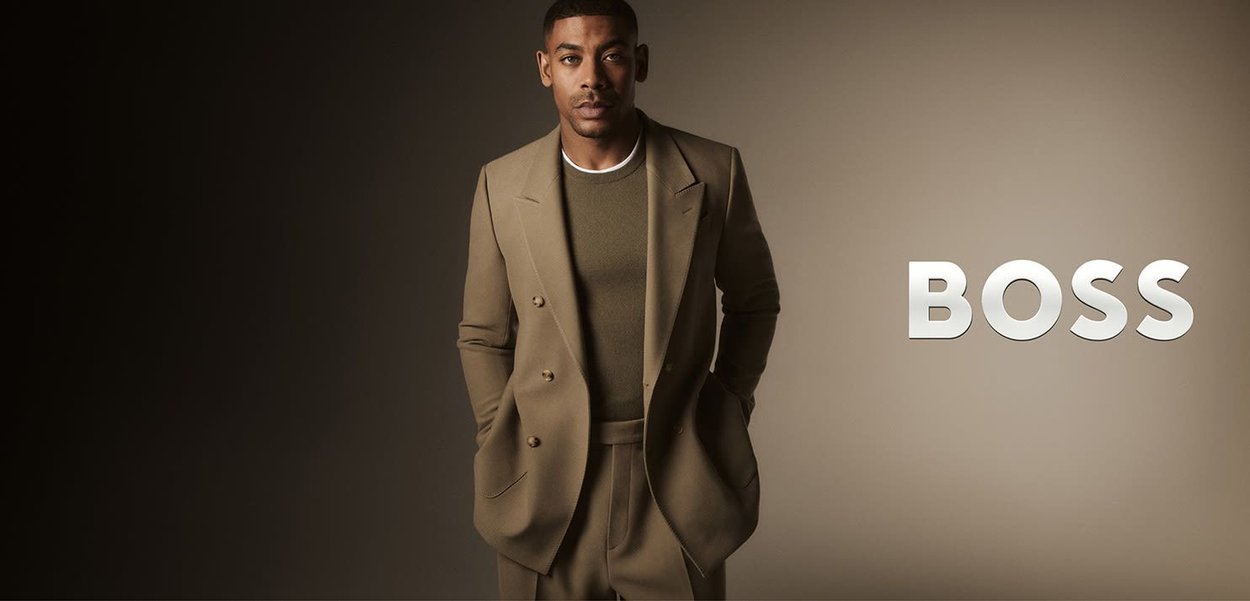 Costume beige sur mesure avec une veste croisée, un pantalon slim et un pull khaki en dessous. Détail du col ras du cou blanc visible.