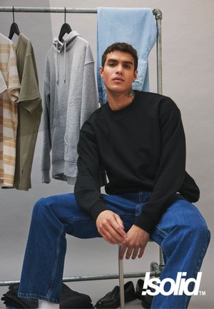 Felpa nera, jeans di denim blu, felpa con cappuccio grigia e maglietta a righe beige-verde su un appendiabiti. La scena mostra un'esposizione di abbigliamento industriale.