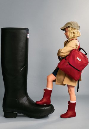 Kind trägt rote Stiefel und beige Mantel, steht mit einem Fuß auf einem übergroßen schwarzen Stiefel und trägt einen roten Rucksack vor einem einfarbigen grauen Hintergrund.