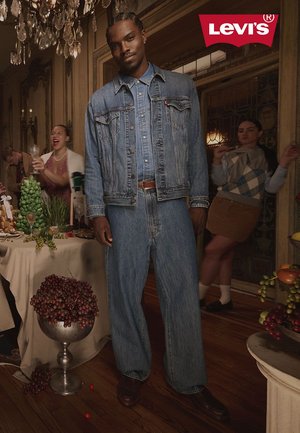 Levi's denim jacket and jeans in light blue, relaxed fit. Wood floor, elegant chandelier, and food table with grapes and vegetables in background.