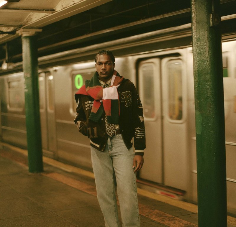 Sort bomberjakke med patches, hvid skjorte, mønstret slips, blå jeans og et farverigt stribet halstørklæde. Metro tog i baggrunden, svagt lys.