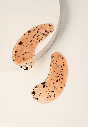 Deux patchs pour les yeux en gel de couleur pêche en forme de croissant avec des specks noirs, à côté d'une plateforme circulaire blanche sur un fond beige.