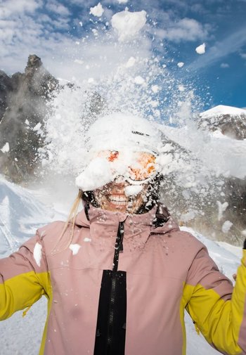 Skijacke in Pink und Gelb, mit schwarzem Reißverschluss. Schneespray umgibt eine lächelnde Person, die einen weißen Helm und orangefarbene Skibrille trägt.