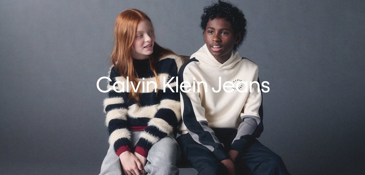 Due bambini siedono fianco a fianco. Uno indossa un maglione a righe bianco e nero e pantaloni grigi, l'altro una felpa crema con accenti scuri.