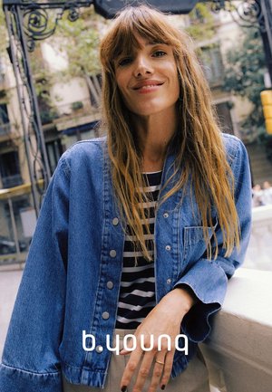 Jeansjacke mit langen Ärmeln, hellblauer Farbe und Druckknöpfen, getragen über einem schwarz-weiß gestreiften Oberteil. Hände ruhen auf einer Kante.