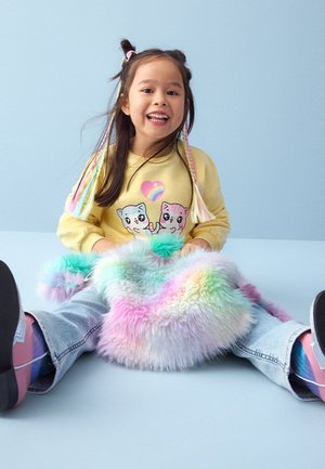 Ragazza giovane con capelli intrecciati colorati che indossa una felpa gialla con gatti, tiene una borsa soffice dai colori pastello, seduta con le gambe aperte.