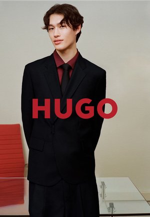 Schwarzer maßgeschneiderter Anzug mit Reverskragen, kombiniert mit einem dunkelroten Hemd und Krawatte. Modell steht vor neutralem Hintergrund, die Arme verschränkt.