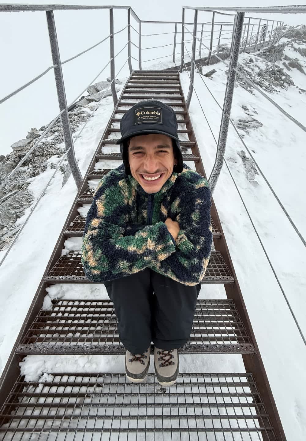 Junge Person in Winterjacke und Mütze sitzt auf verschneiten Metalltreppen mit Geländern auf einem Bergweg, lächelt und hat die Arme verschränkt.