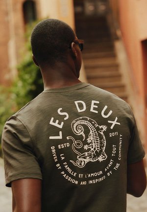 Man draagt een olijfgroen t-shirt met "LES DEUX" en een hagedisafbeelding op de rug, wandelend buiten nabij trappen.