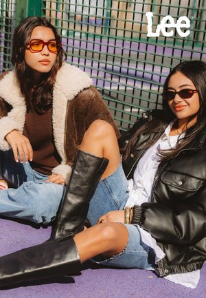 Due modelli seduti su una superficie viola. Uno indossa un giubbotto di shearling marrone e occhiali da sole arancioni. L'altro indossa un giubbotto di pelle nera e stivali neri alti.