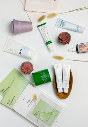 Neuf produits de soin de la peau comprenant des tubes de crème, un pot de crème hydratante et des masques en feuille de différentes couleurs (blanc, vert, bleu clair et violet) sur une surface blanche.