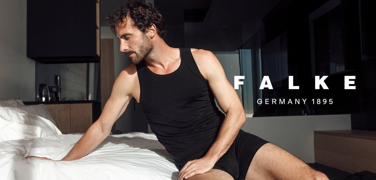 Black tank top and shorts worn by a man, seated on a bed with white bedding, in a modern room with wooden furniture and natural light.