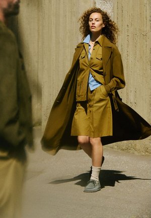 Olive green trench coat over a light blue shirt and matching skirt. Model wears gray shoes and white socks, standing against a textured wall.