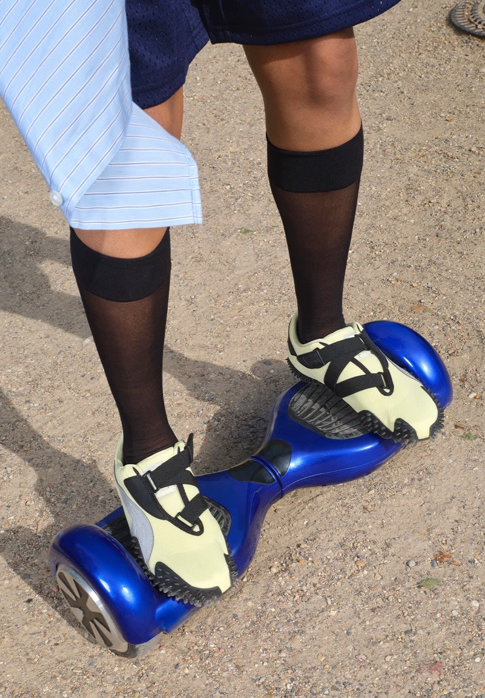 Blue hoverboard with black wheels beneath a person wearing yellow and black shoes along with black knee-high socks. The ground is sandy.
