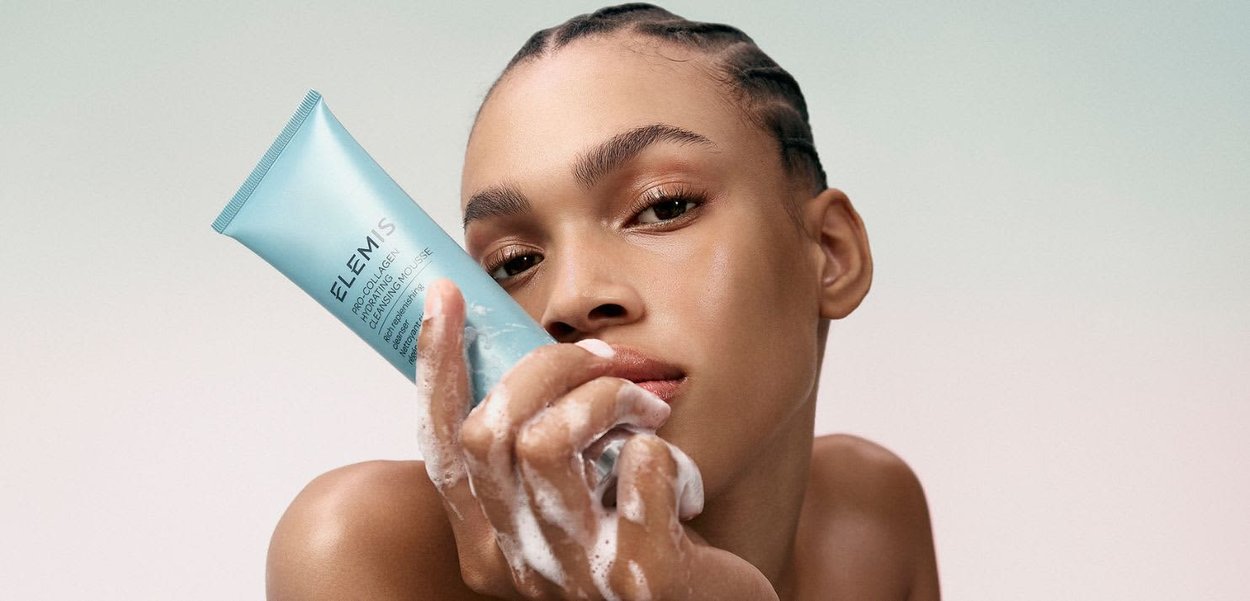 Person med flette holder et skumrensemiddelrør merket "Elemis Pro-Collagen Hydrating Cleansing Mousse" nær ansiktet.