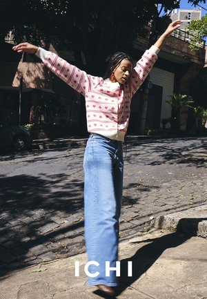 Frau, die draußen auf einer sonnigen Straße tanzt, trägt einen rosa gemusterten Pullover und weite blaue Jeans mit Ohrhörern im Ohr.