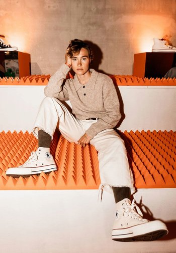 White high-top sneakers with black accents, beige sweater, and white jeans, seated on textured orange backdrop with a concrete wall.