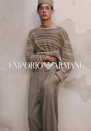 Hombre joven con suéter beige estampado, sombrero a juego y pantalones grises de rayas de talle alto, de pie contra una pared texturizada.