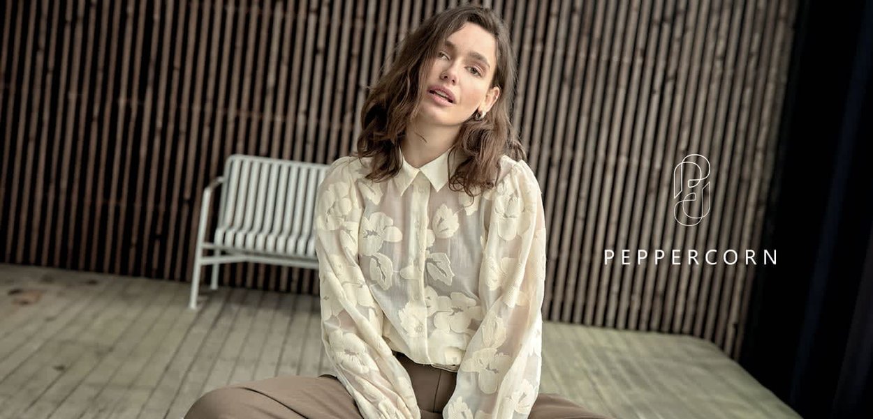 Femme aux cheveux bruns ondulés portant un chemisier blanc transparent à fleurs et un pantalon marron, assise sur une terrasse en bois avec un banc blanc derrière elle.
