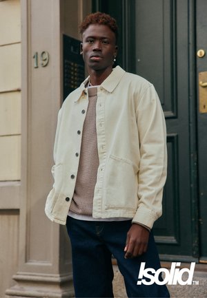 Giacca in tela beige chiaro con bottoni scuri, indossata sopra un maglione beige lavorato a trama. Abbinata a jeans blu scuro. Sullo sfondo una porta.