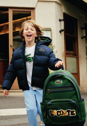 Giacca a vento blu navy, dettagli verdi, camicia bianca con logo, pantaloni azzurri chiari, zaino verde con ricamo "LACOSTE" e dettagli del logo.