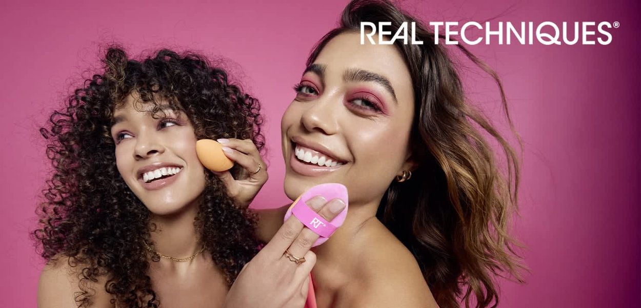 To kvinder med forskellig hårtekstur holder makeup-svampe: en orange og en pink med "RT"-logo. Den lyse pink baggrund fremhæver de levende farver.