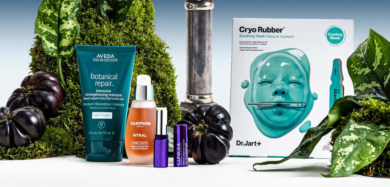 Tubetto verde di Aveda, siero Darphin in bottiglia ambra, flacone viola Clinique, confezione maschera Dr. Jart+ turchese con maschera a forma di volto, circondati da melanzane nere e foglie.