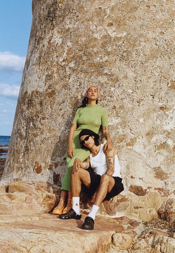 Due persone posano su un terreno roccioso vicino a una struttura in pietra texture. Una indossa un vestito verde a costine e scarpe con platform arancioni; l'altra, una canotta bianca, pantaloni corti neri e crocs neri.