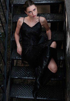 Schwarzes Paillettenkleid mit Spaghettiträgern, figurbetonter Silhouette und strukturiertem Muster. Das Model trägt knielange schwarze Lederstiefel auf metallischen Treppen.