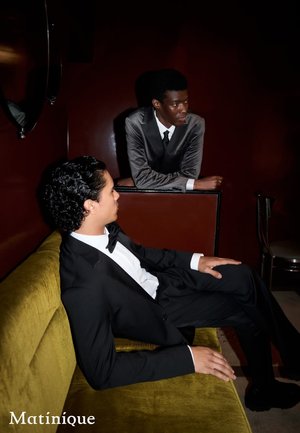 Zwei Männer in formeller Kleidung, einer in einem schwarzen Anzug mit Fliege, sitzen auf einem strukturierten olivgrünen Sofa; der andere in einem grauen Anzug lehnt sich an eine dunkle Oberfläche.