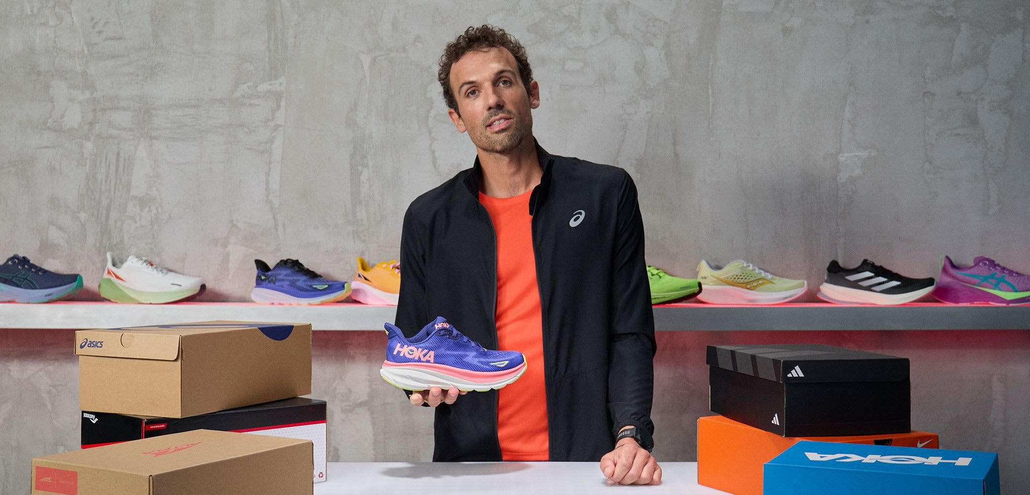 HOKA Sneaker in Blau mit pinken Akzenten, ausgestattet mit einer dicken weißen Sohle. Nebenbei sind verschiedene Schuhkartons gestapelt, im Hintergrund sind bunte Sneaker zu sehen.