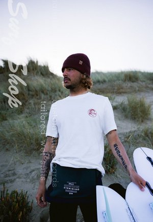 Witte t-shirt met rood logo, donkere beanie en zwarte utility tas. Surfboard deels zichtbaar, staande in het gras met een zandige achtergrond.