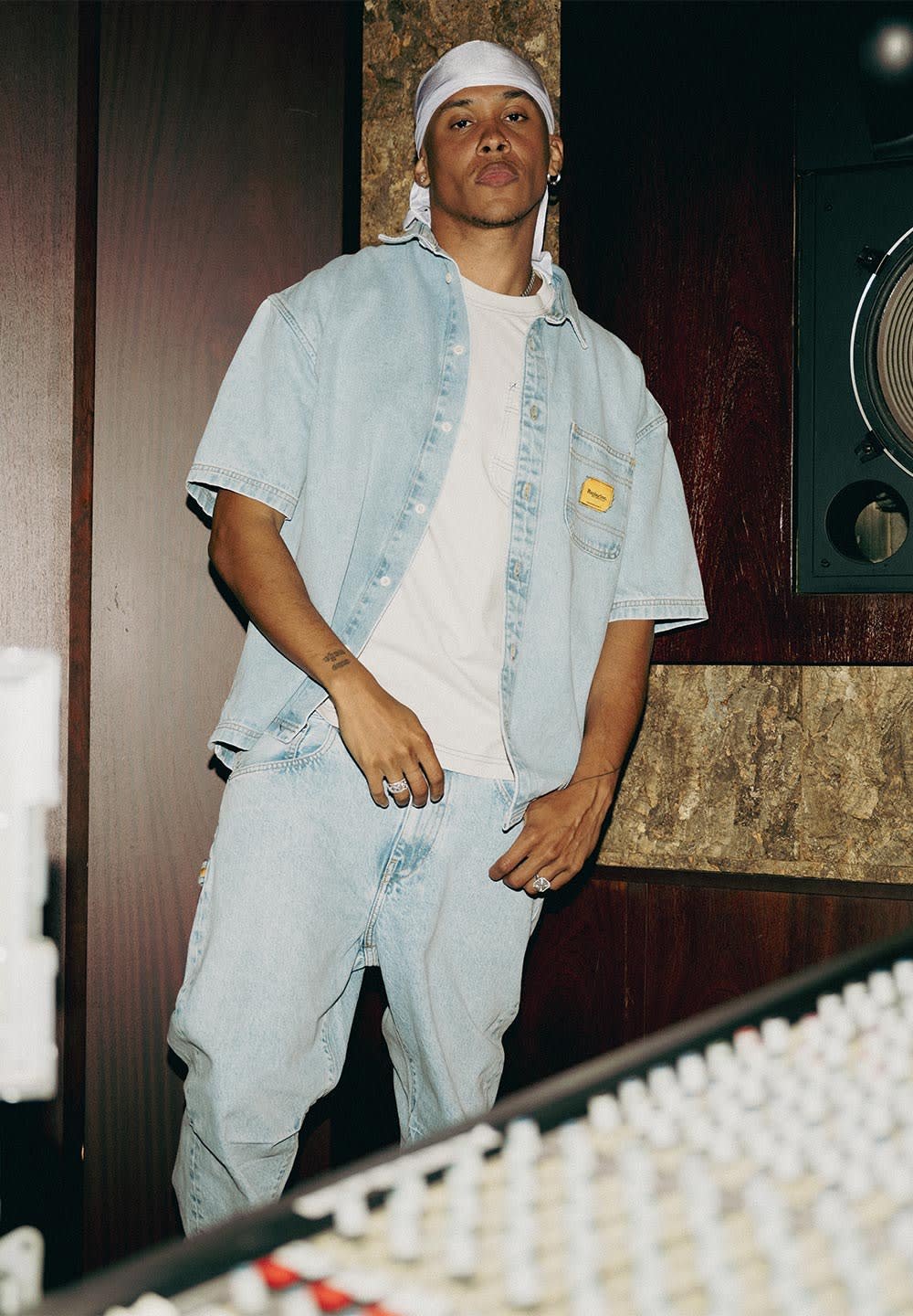 Mann in einem hellen Jeanshemd und Jeans mit einem weißen Bandana steht in einem Tonstudio neben einem Mischpult und einem Lautsprecher.