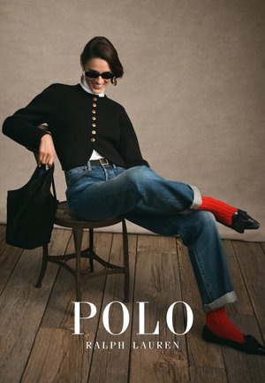 Mustat lyhyet neuleet kultaisilla napilla, valkoinen korkea kaulus, siniset farkut ja punaiset villasukat, yhdistettynä mustaan laukkuun ja istuen tuolilla.